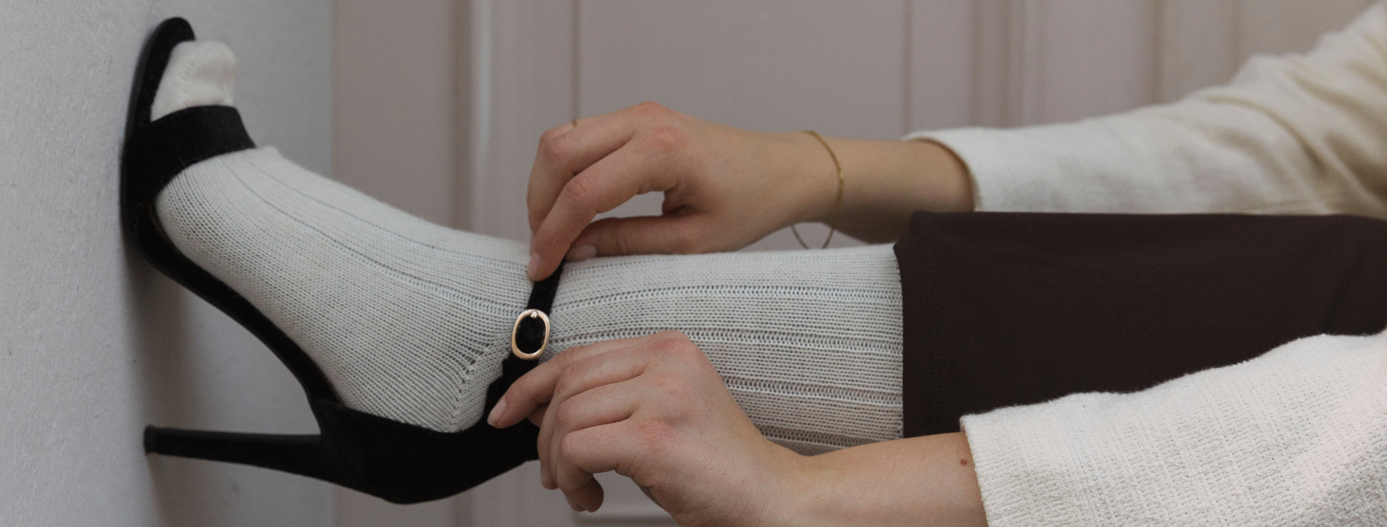 Person adjusting a black high-heeled shoe with white socks against a neutral background